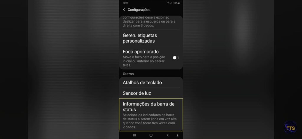Descrição da imagem:
No topo aparece “Configurações” e, na barra de status, o horário 18:11 e bateria em 43%.

Itens visíveis na lista:
Geren. etiquetas personalizadas
Foco aprimorado (com uma chave/toggle à direita, aparentemente desligada)
Outros
Atalhos de teclado
Sensor de luz
Informações da barra de status (este item está destacado com uma borda amarela)

Texto pequeno abaixo de “Informações da barra de status”:
“Selecione os indicadores da barra de status a serem lidos em voz alta quando você tocar três vezes com 2 dedos.”

No canto inferior direito há um pequeno logotipo circular com as letras “TS”. Fim da descrição 